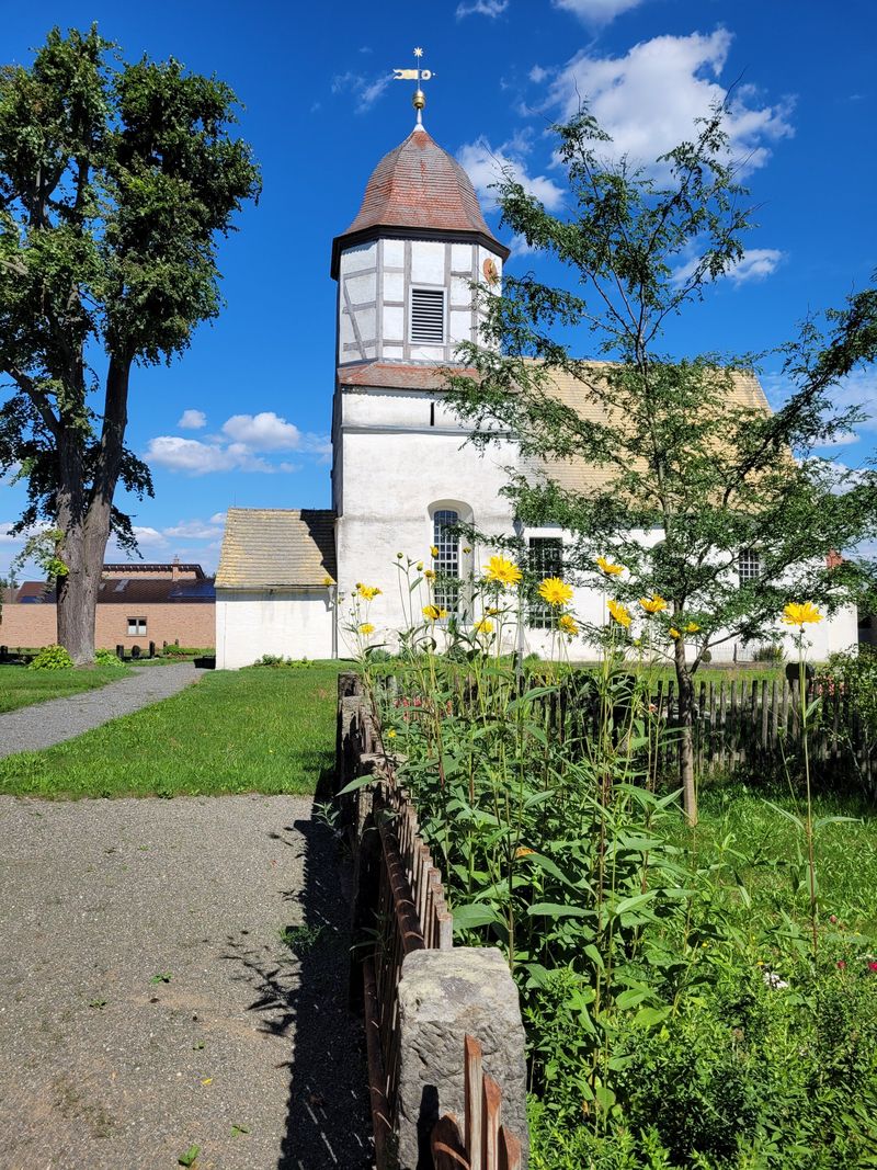 Weiße Dorfkirche mit Fachwerkturm, davor gelb blühende Sonnenblumen