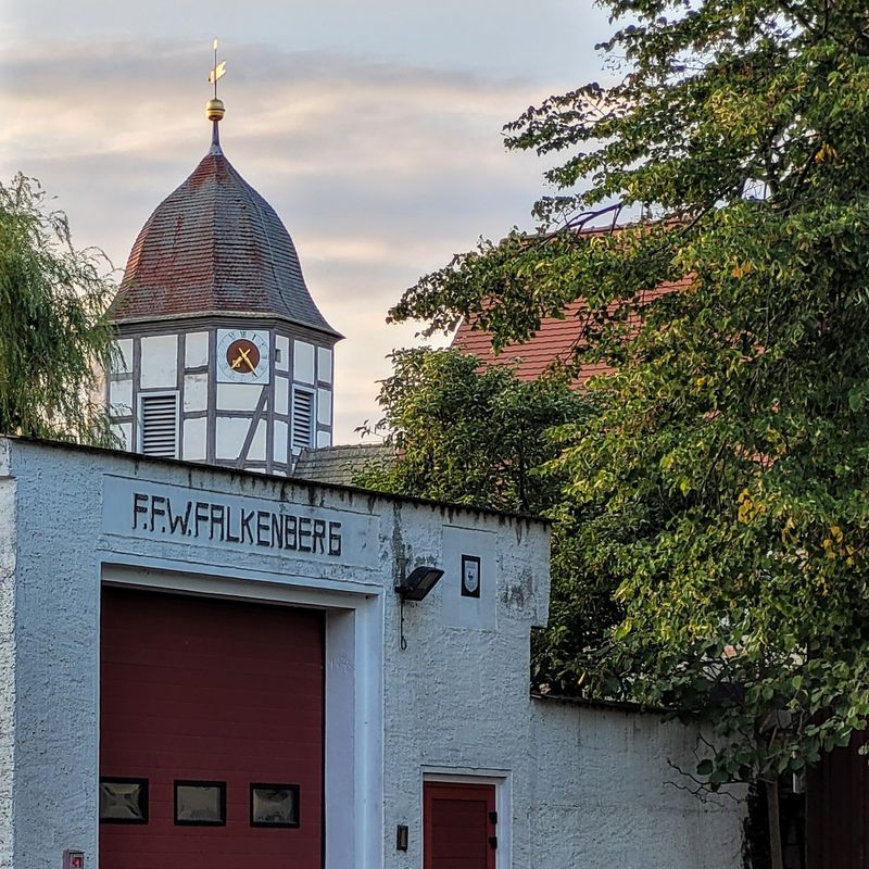 Feuerwehrhaus mit Aufschrift F.F.W. Falkenberg, dahinter ein Fachwerk-Kirchturm